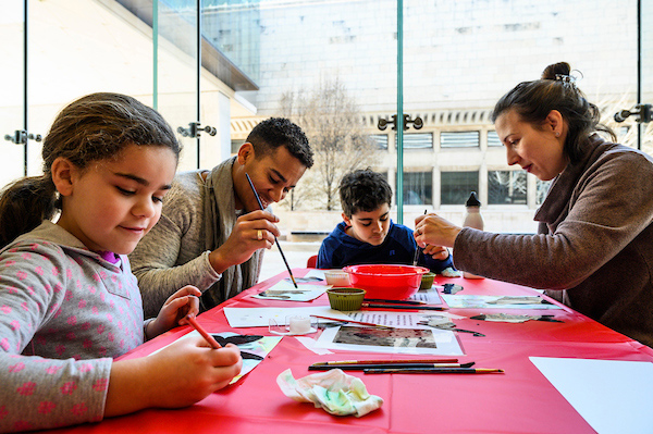 A family of four sits together at a table covered with a red tablecloth. Each person is working on a painting.