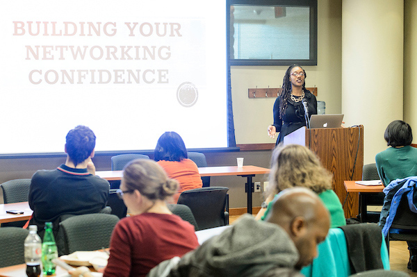 A woman stands behind a podium next to a powerpoint presentation, while a group of students listen and take notes.