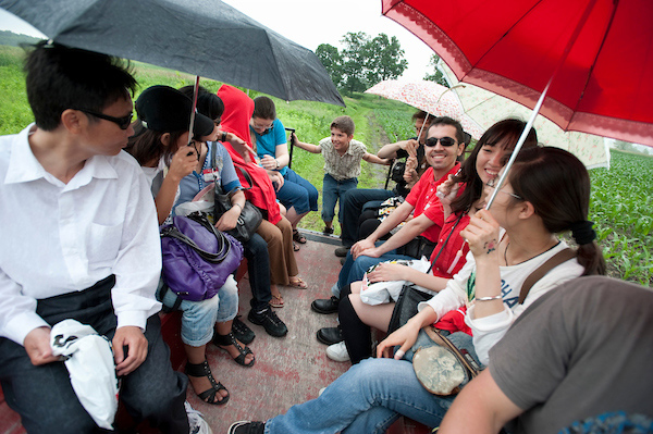 Smiling students sit together in a wagon, some holding umbrellas.