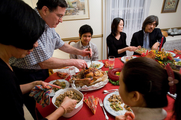 People sit together around a table draped with a red tablecloth while one person carves a turkey on the table.