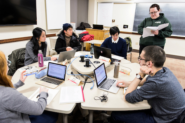 Students cluster around a table with open laptops while a graduate student stands and leads a discussion.