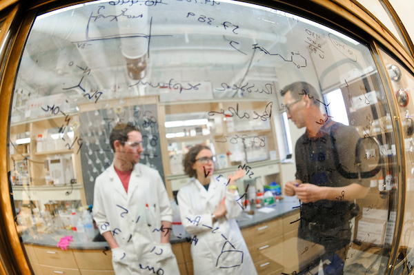 A man wearing a black coat and two students in white coats stand behind a see-through glass board with chemistry notations.