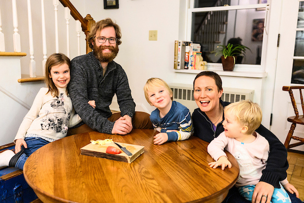 Two adults and three children sit around a table in their home, smiling for the camera.