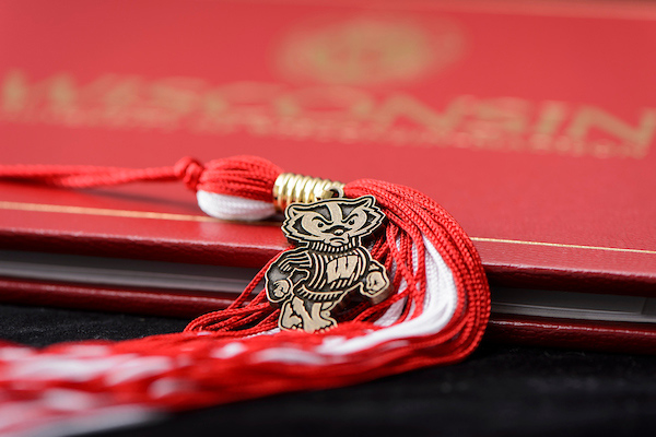 A Bucky Badger pendant rests on a red and white graduation tassle, with a Wisconsin diploma cover in the background.