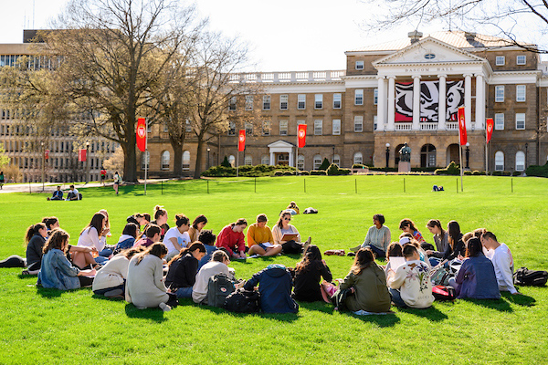A circle of students sit on Bascom Hill with the Bucky banner on Bascom Hall in the background.