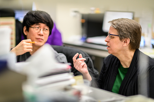 Two people seated at a desk with computers converse with one another.