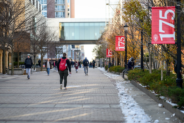People wearing backpacks walk down a stone pedestrian mall with red banners visible along the sides.
