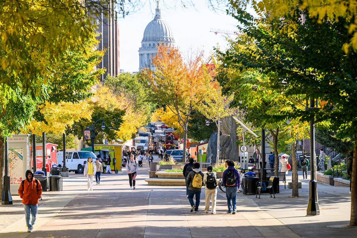 People walk on a pedestrian mall underneath green and yellow trees. The white dome of the state capitol building is visible in the background.