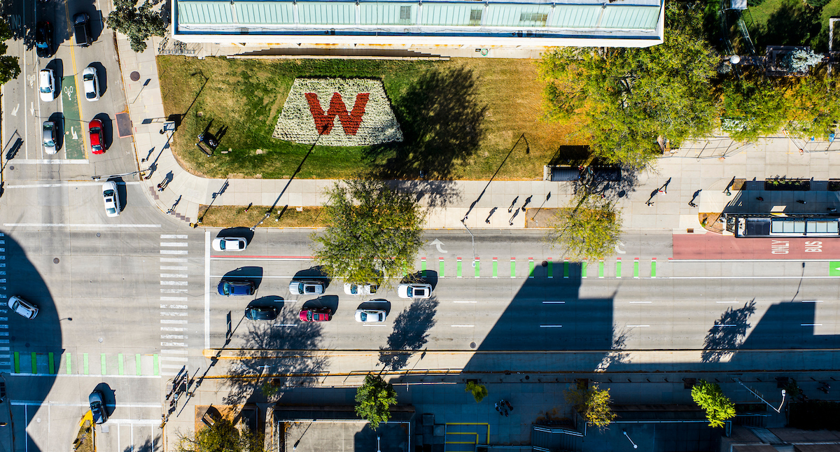 Aerial view of a wide street with cars and a bus on it. A flowerbed with a decorative red W made of plantings is visible.